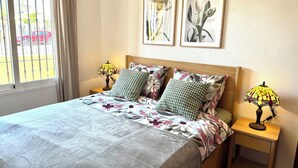 Cama bem arrumada com roupa de cama floral, duas luminárias de cabeceira e dois quadros decorativos com flores na parede.