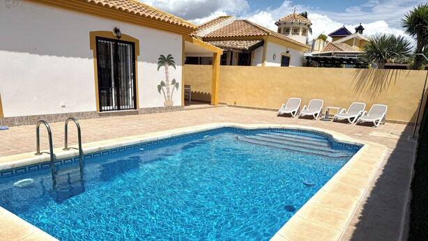Uma piscina retangular com água azul cristalina, rodeada por um deck ladrilhado. Há espreguiçadeiras e uma pequena área de pátio com mesa e cadeiras.