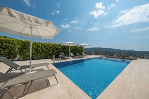 Ein Swimmingpool mit Liegestühlen und Sonnenschirmen, umgeben von einer Hecke und Blick auf ferne Hügel.