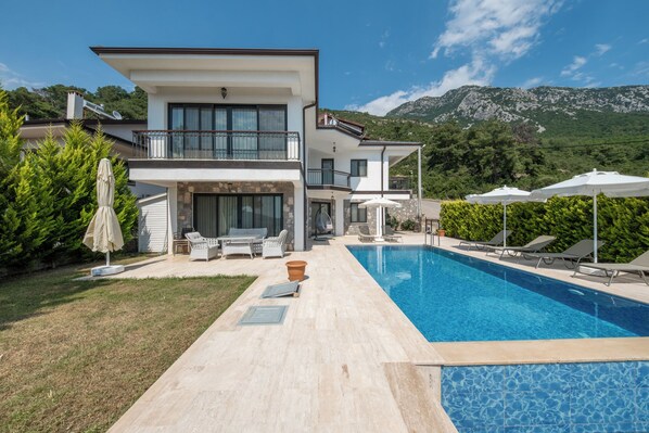 A modern two-story house with a swimming pool, outdoor furniture, and a view of mountains.
