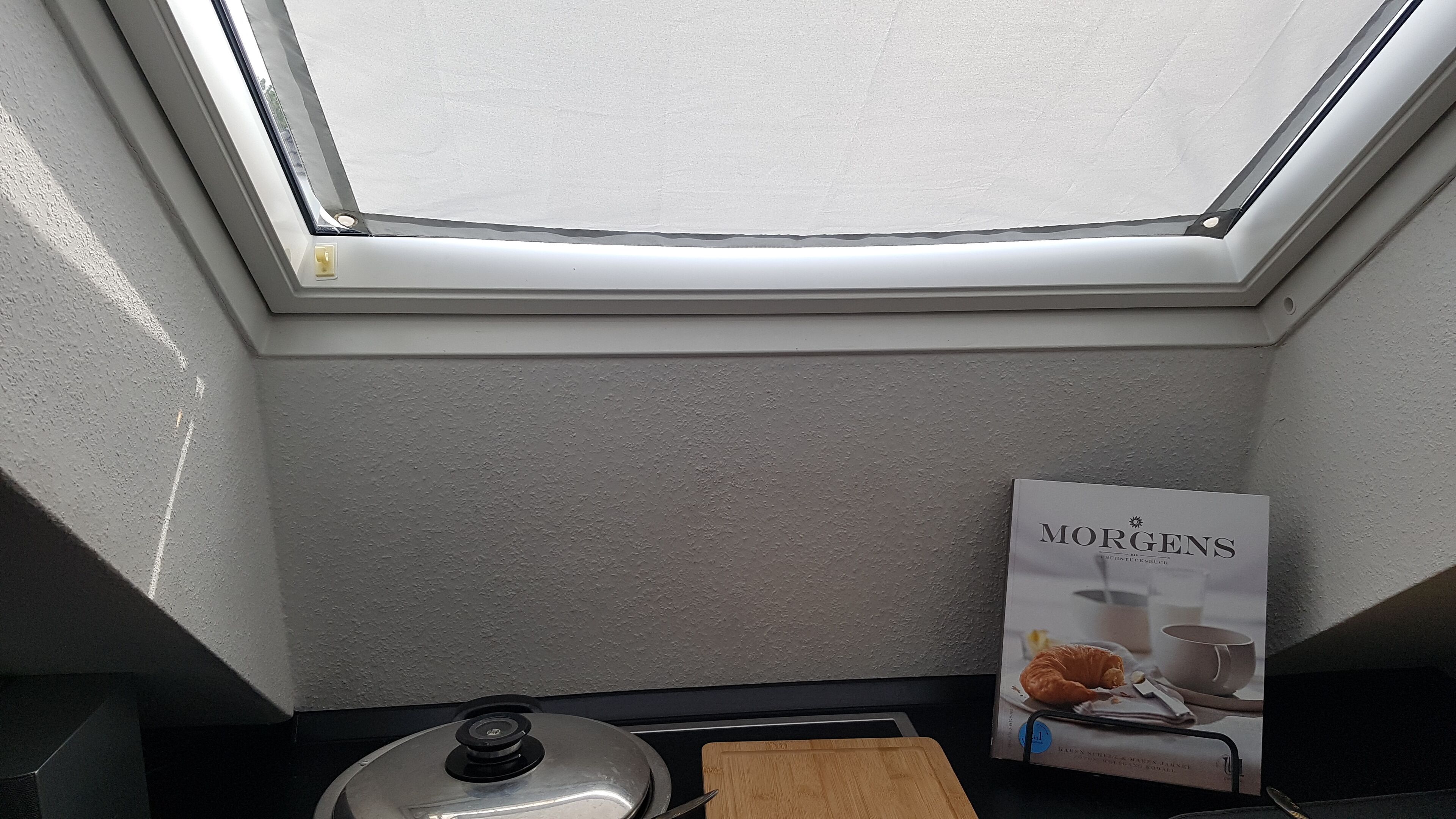 Une cuisine avec un puits de lumière, une planche à découper en bois, une casserole en métal et un magazine MORGENS posé sur le comptoir.