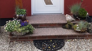 A red building with a white door featuring a unique geometric design. There are potted plants and a small garden area in front of the door. - Apartment (Brunsbüttel)