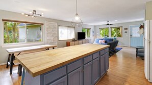 Eine geräumige Küche mit einer großen Kochinsel, Holz Arbeitsplatten und grauen Schränken. Es gibt einen Essbereich mit Tisch und Stühlen, sowie ein Wohnzimmer mit Sofa und Fernseher.