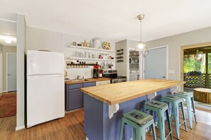 Uma cozinha com ilha central, banquetas, geladeira e diversos utensílios em prateleiras.