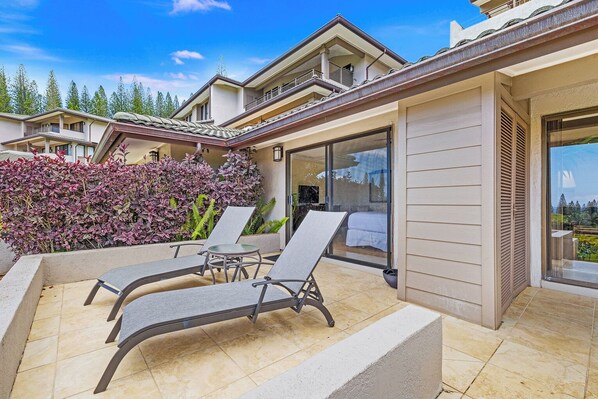 A modern outdoor patio with two lounge chairs and a small table, surrounded by a low hedge and a large window offering a view of the surrounding area.