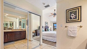Una habitación de hotel con una puerta corrediza de vidrio que da a un baño, una cama con cabecera, un ventilador de techo y un cuadro en la pared.