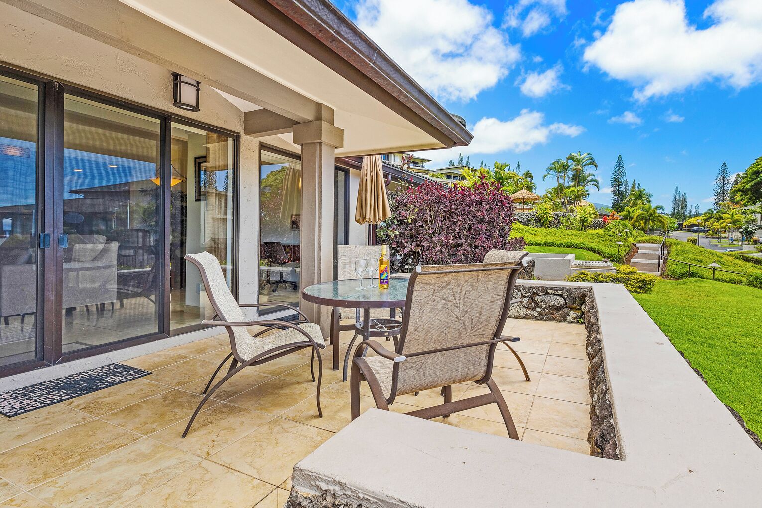 Une terrasse dotée d’une porte vitrée donnant sur un espace extérieur abrité, aménagé avec deux chaises et une table, ainsi qu’un petit jardin avec un bac en pierre et des plantes tropicales.