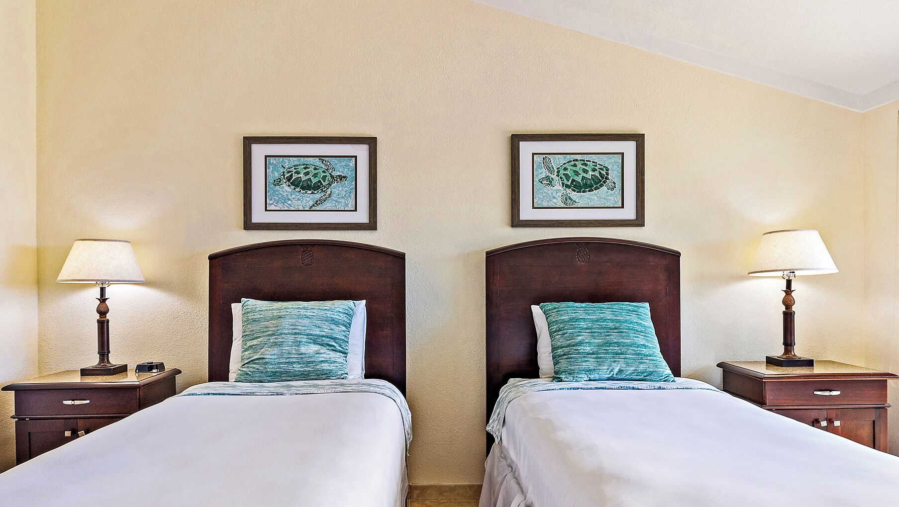Een hotelkamer met twee bedden, elk met een houten hoofdbord en een bijpassend nachtkastje. Boven de bedden hangen twee ingelijste foto's, en op de nachtkastjes staan twee lampen.