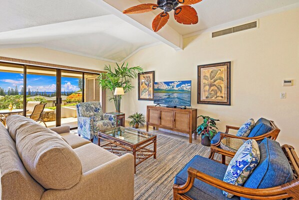 A spacious living room with a large sofa, a coffee table, and two armchairs. There is a television on a wooden cabinet, and a ceiling fan above.