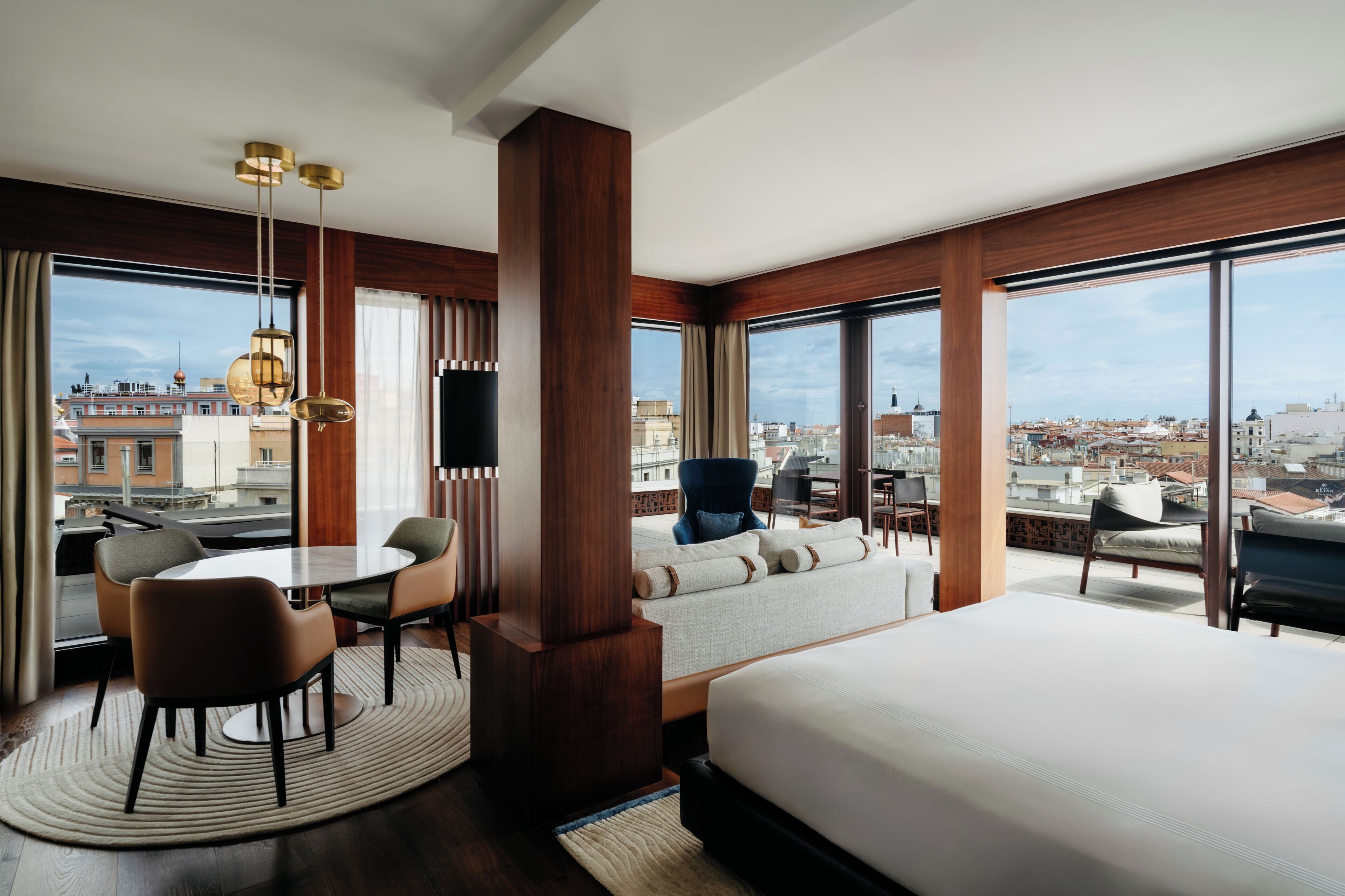 A modern hotel room with a large bed, a dining area with chairs, a sofa, and a view of the cityscape through large windows.