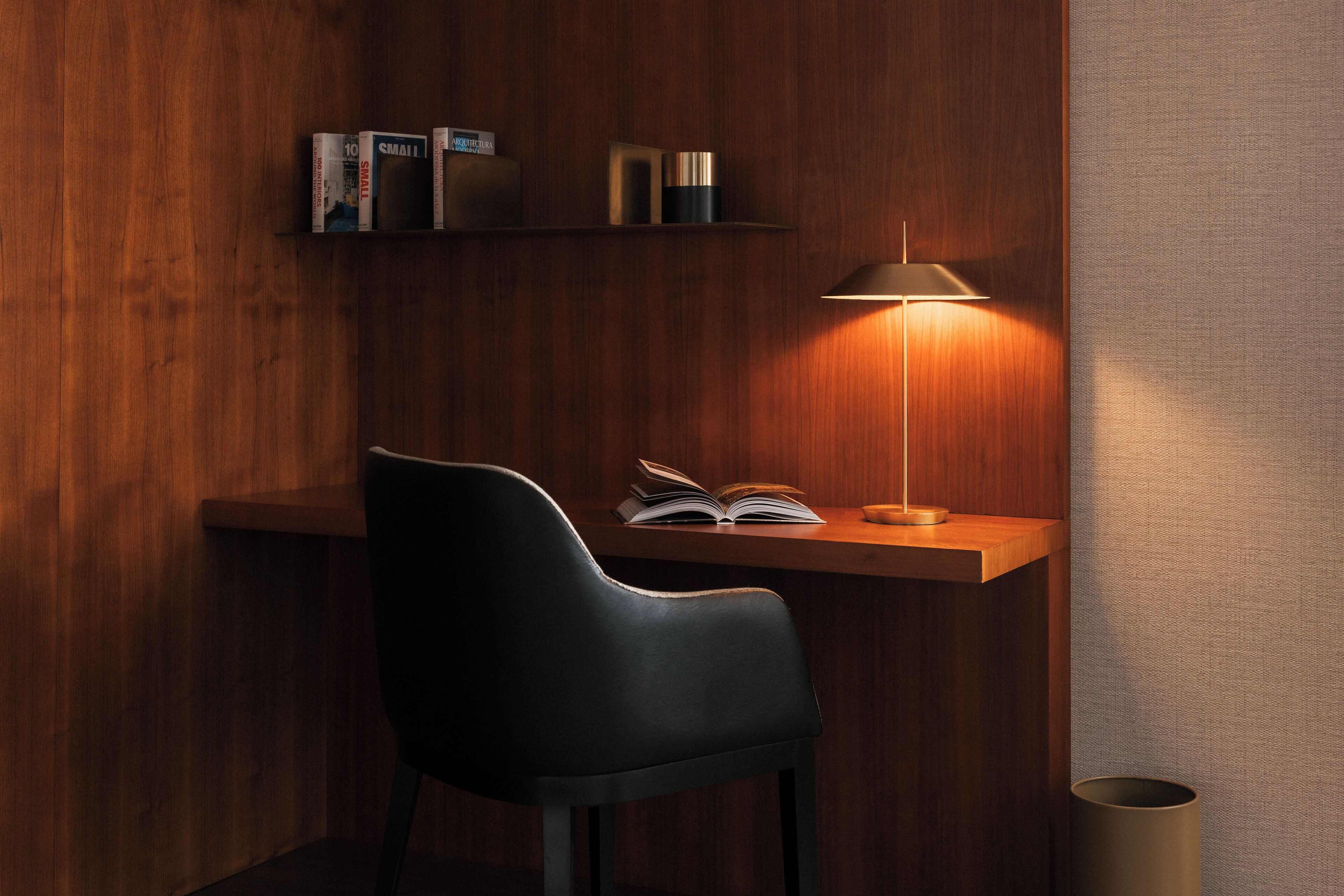 A wooden desk with a lamp, a chair, and a shelf with books and decorative items.
