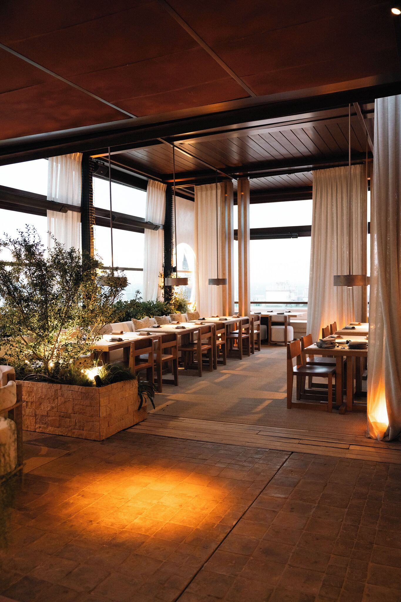 A restaurant with a long dining area, wooden tables, and chairs. There are large windows with curtains, and a view of the city.