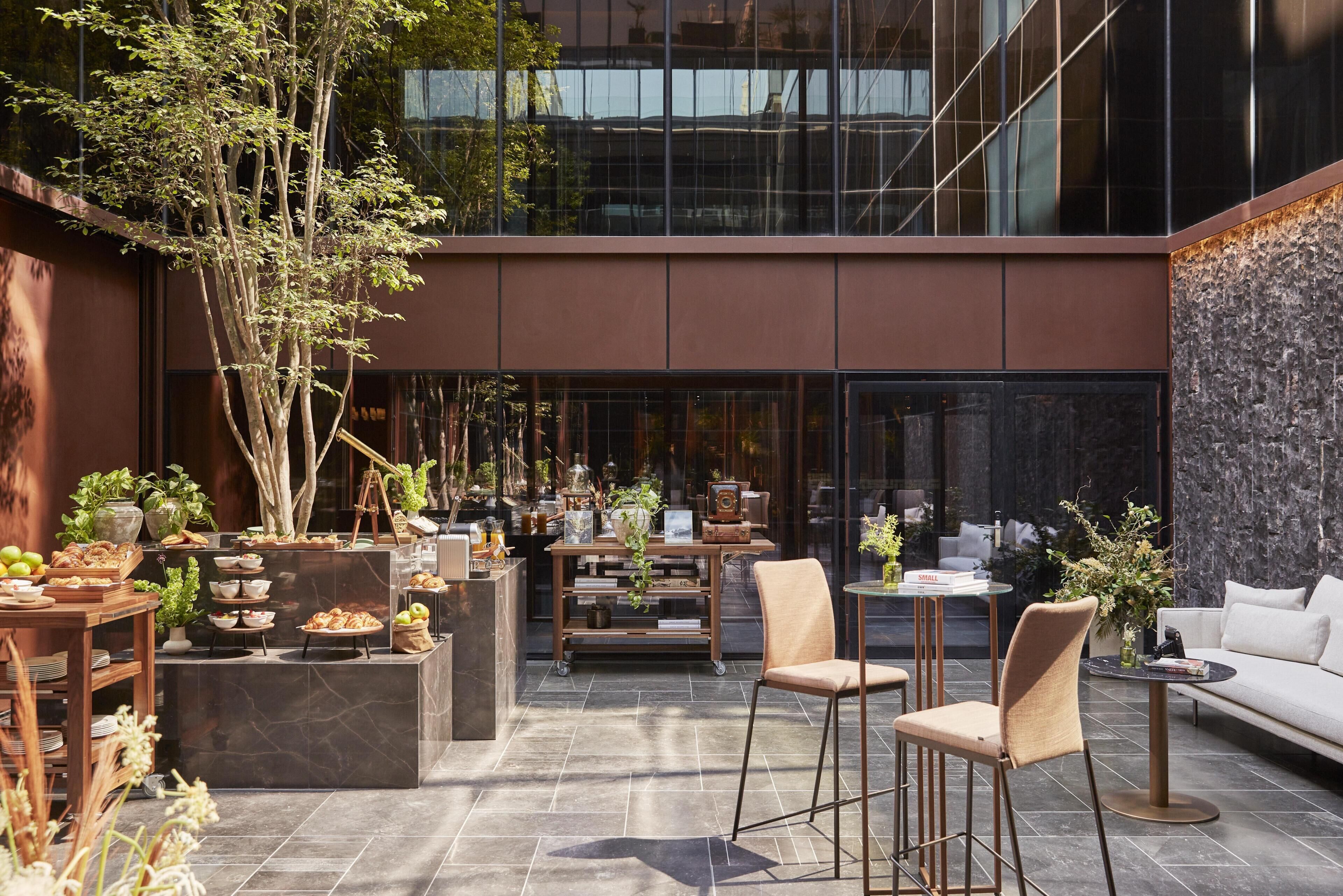 Un espacio moderno para comer al aire libre con diversas opciones de asientos, una mesa central con un florero y un edificio de fondo con grandes ventanales.