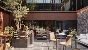 Un espacio moderno para comer al aire libre con diversas opciones de asientos, una mesa central con un florero y un edificio de fondo con grandes ventanales.