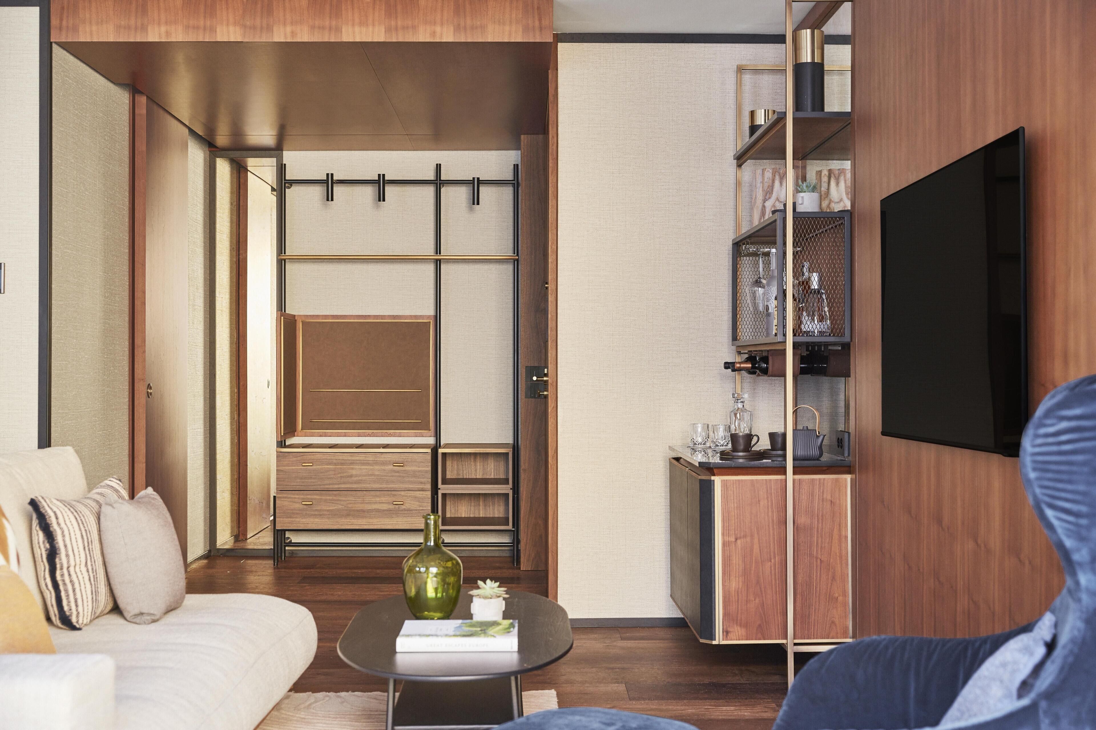Una sala moderna con sofá, mesa de centro y televisor. Se observa un mueble de madera y una pequeña zona de cocina con estantería.