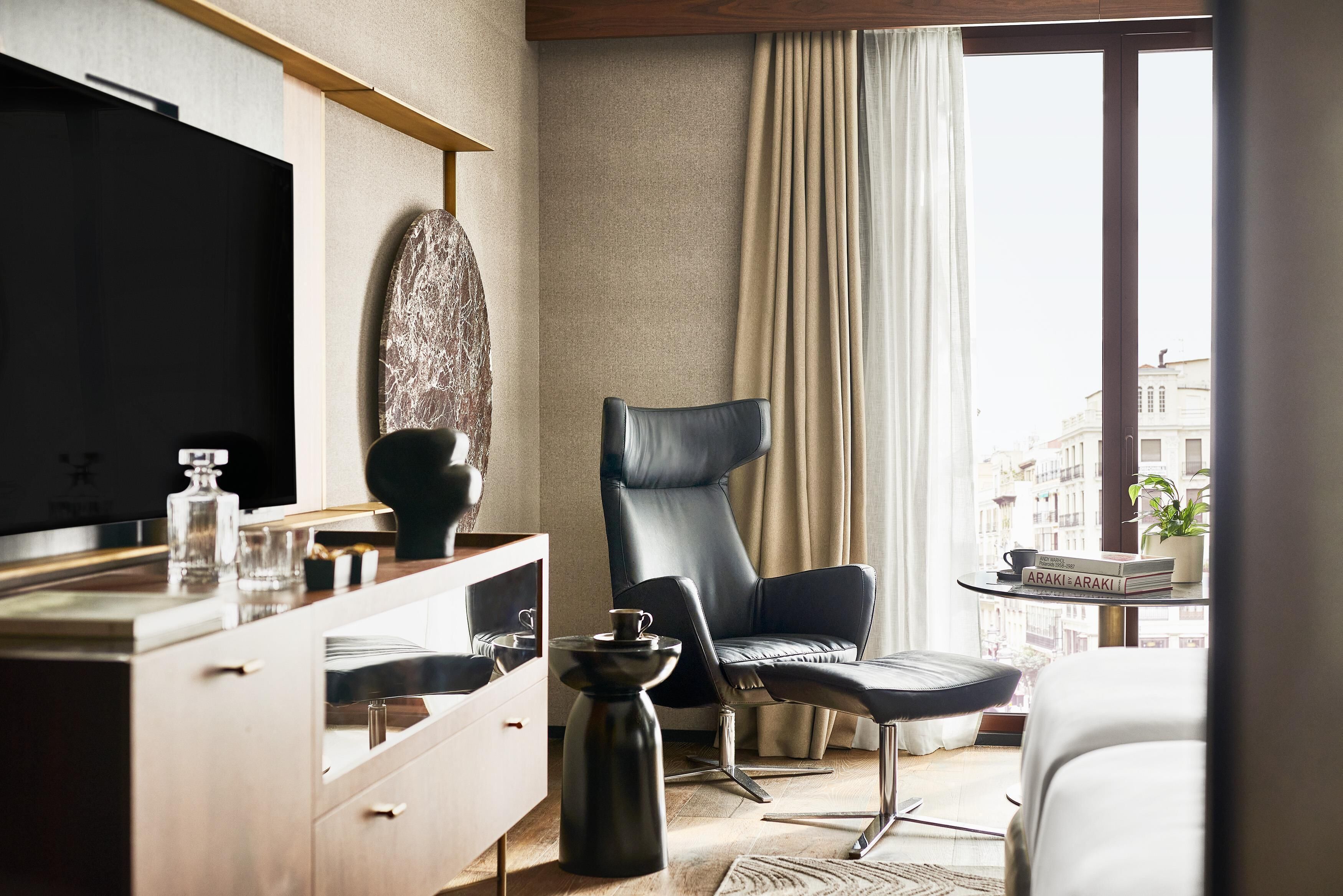 A modern hotel room with a large flat-screen TV, a leather armchair, a small round table with a coffee pot, and a view of the city through large windows.