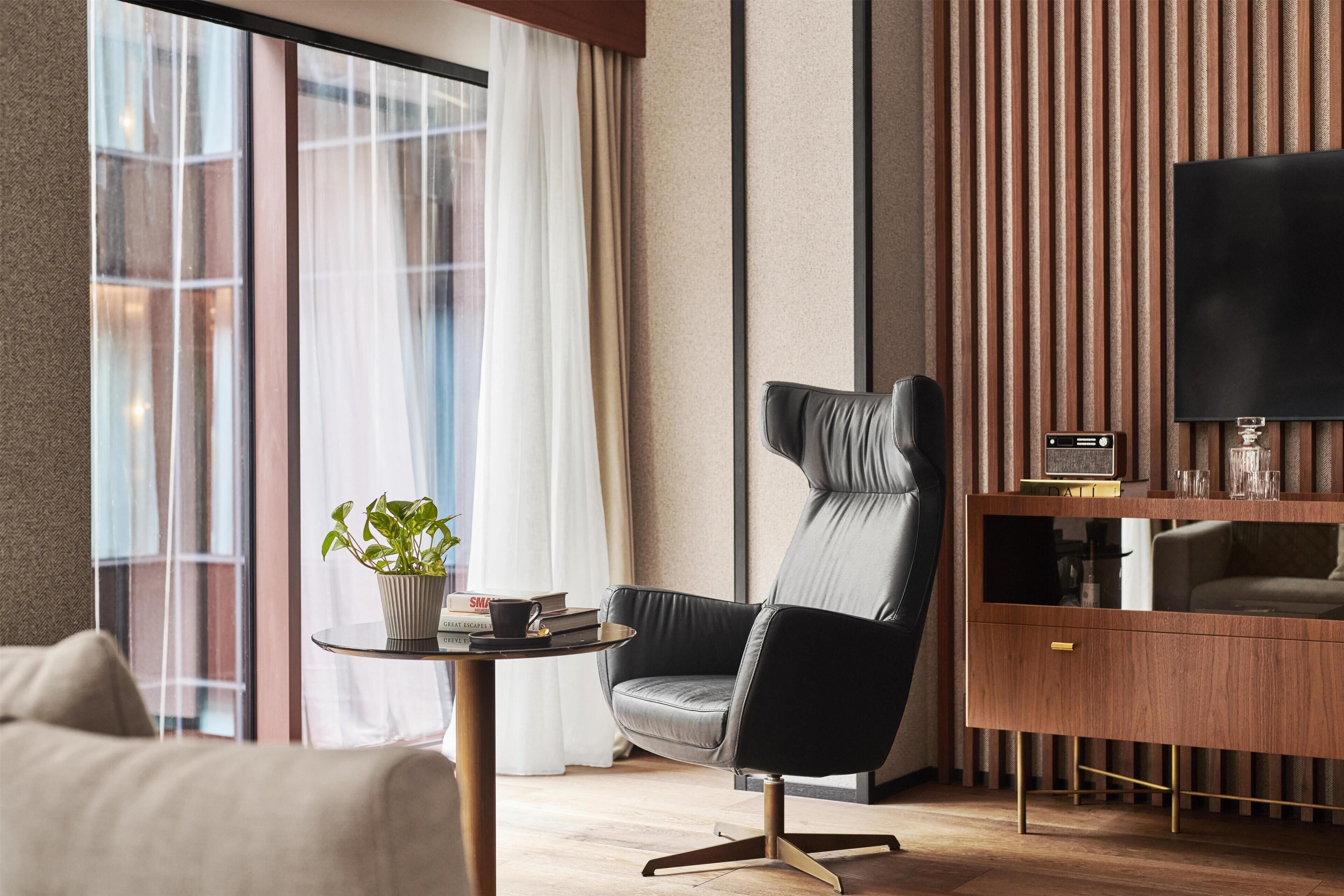 Una habitación de hotel moderna con un sillón de cuero, una mesita con una planta, un televisor de pantalla plana y una consola de madera.
