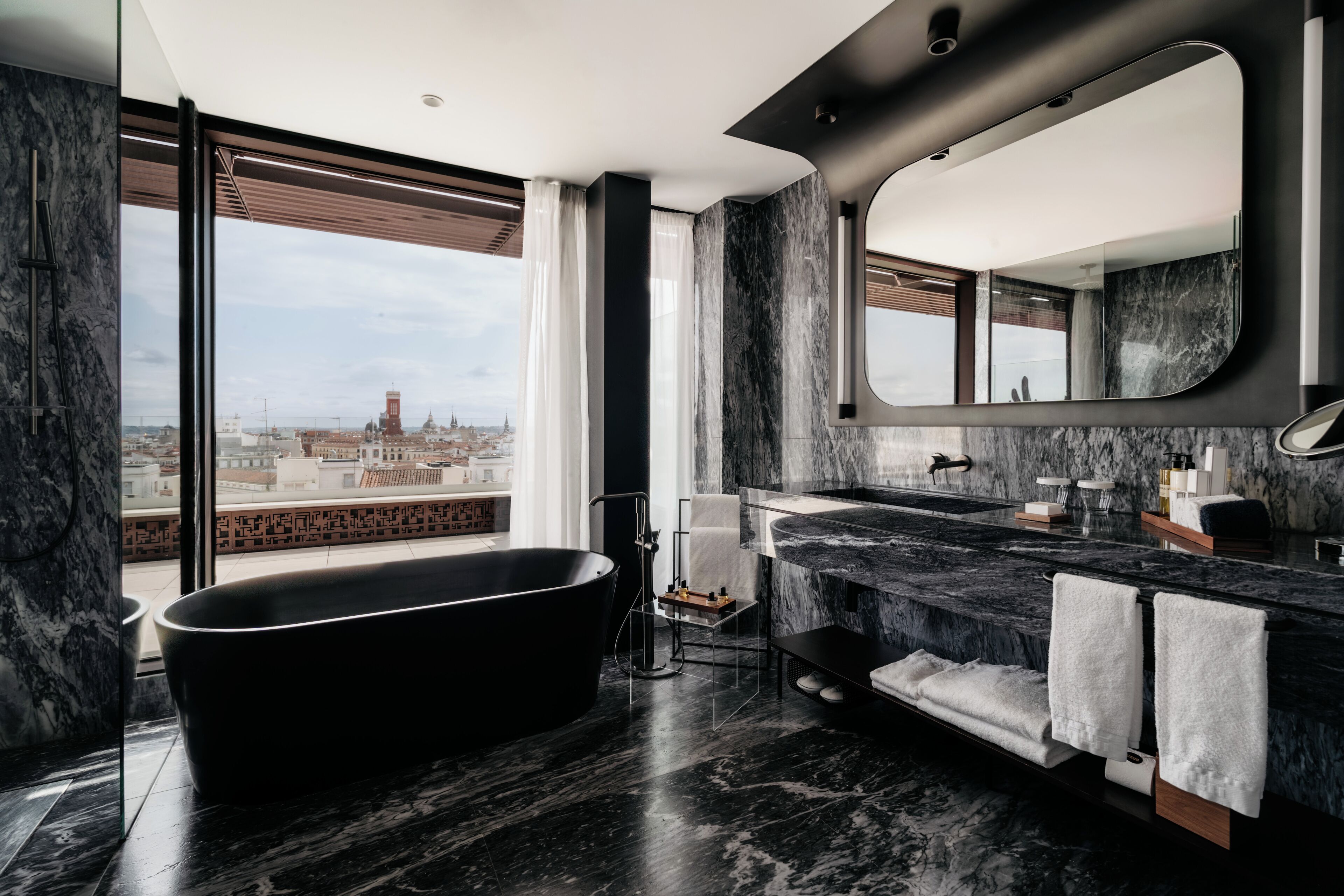Un baño moderno con una bañera independiente, un espejo grande y vistas a un paisaje urbano a través de amplios ventanales.