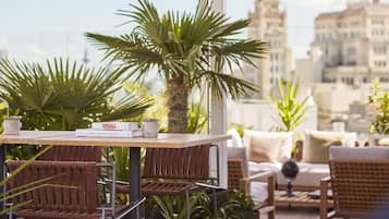 Una terraza con mobiliario de mimbre, una mesa con libros y vistas a la ciudad.