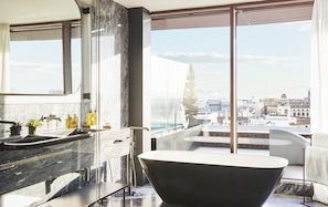 A modern bathroom with a large bathtub, a sink with marble countertops, and a city view through large windows.