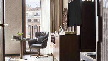 Una habitación moderna con un sillón de cuero, una mesita con una radio y un libro, un escritorio con un recipiente de vidrio y un ventanal con vistas a edificios.