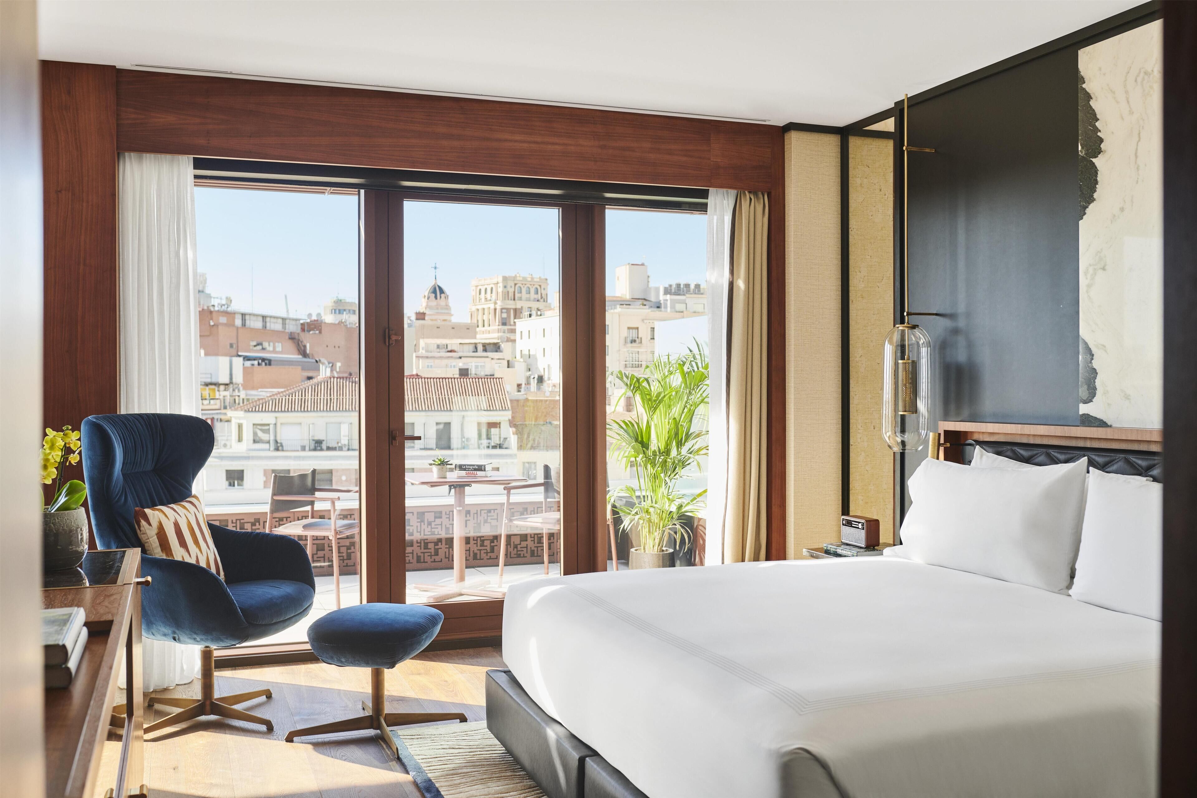 Una habitación de hotel con una cama grande, un sillón azul, una mesita pequeña y vistas de la ciudad a través de amplios ventanales.