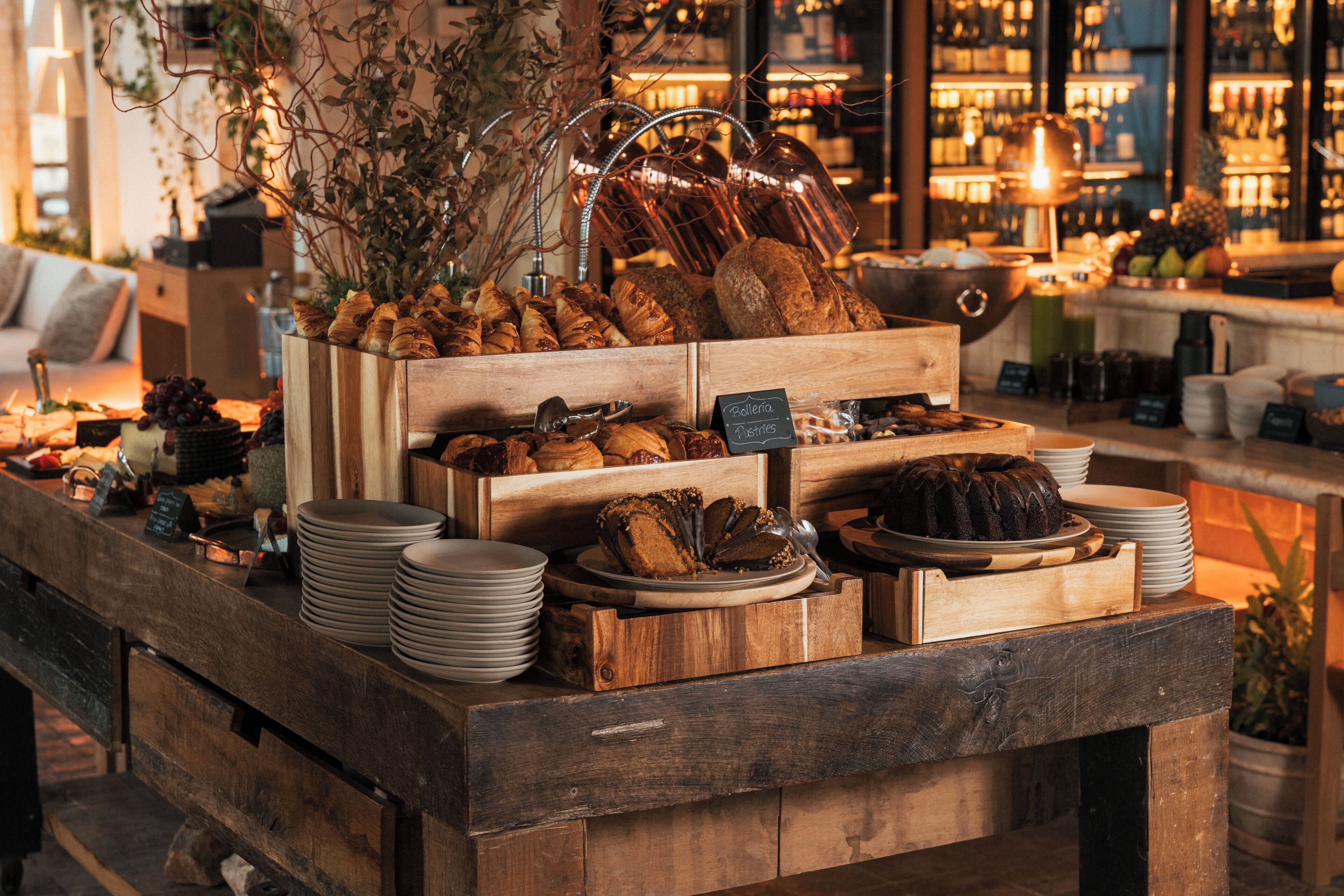 A wooden buffet table with various baked goods and pastries, surrounded by a bar area with shelves of bottles.