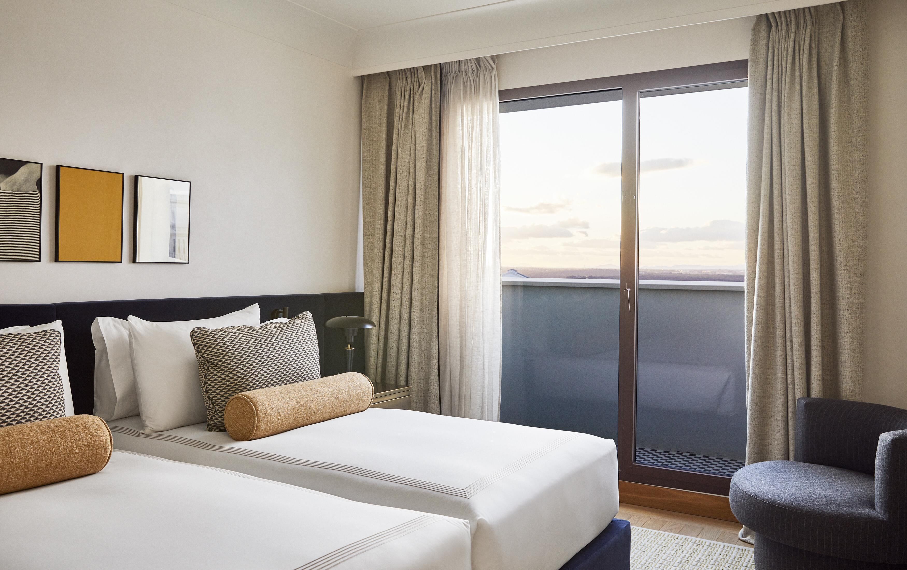 A hotel room with a bed, a chair, a nightstand, and a view of the city.