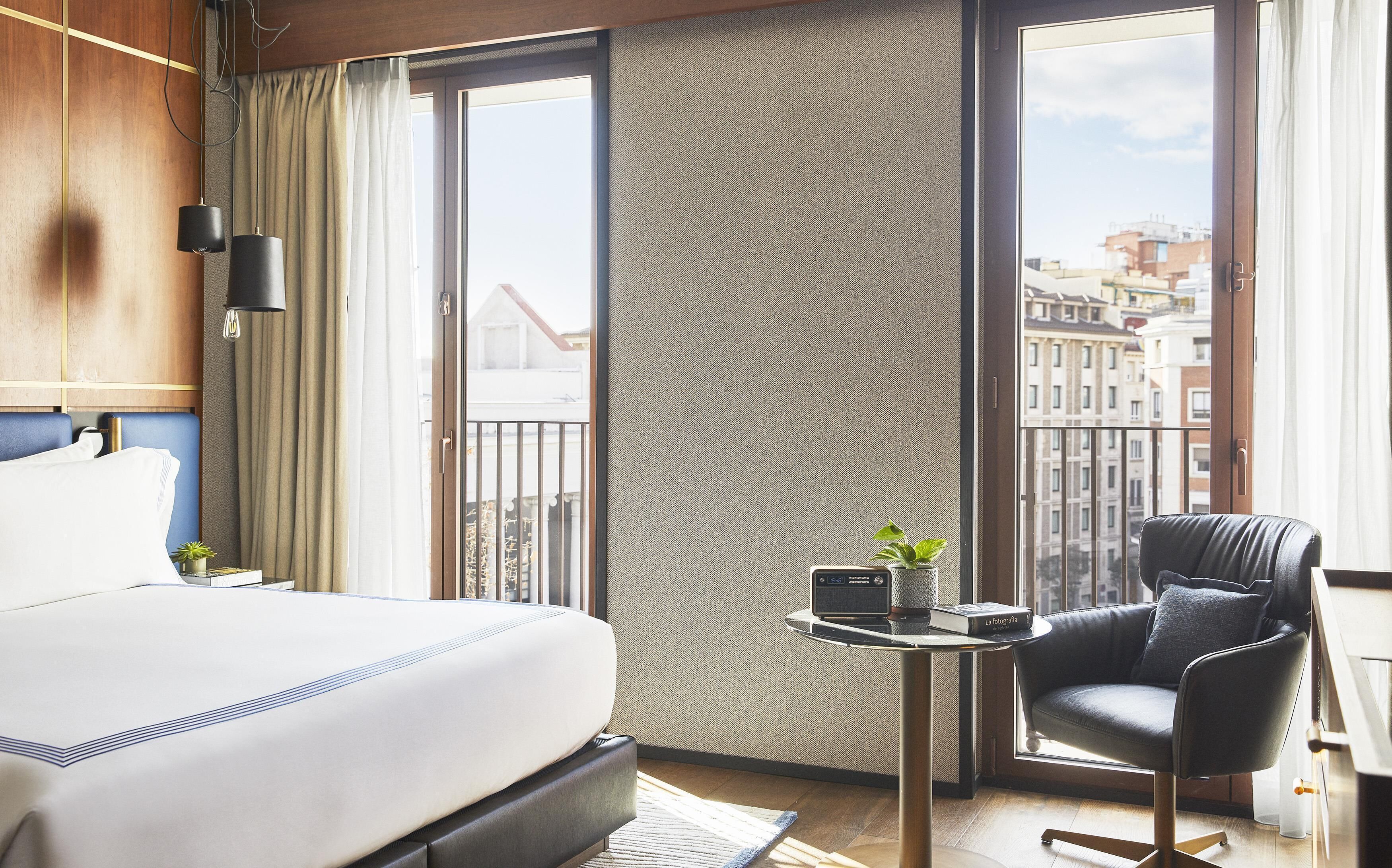 Una habitació d'hotel amb un llit, una cadira, una taula petita amb una planta i un llibre, i una vista de la ciutat a través de grans finestres.