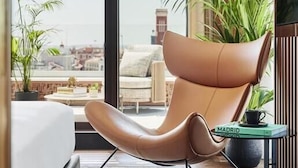 A modern hotel room with a large bed, a leather armchair, a small table, and a view of the city. - Thompson Madrid, by Hyatt (Madrid)