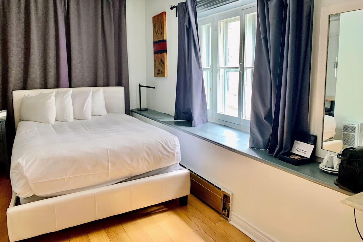 A bedroom with a bed, a window with curtains, a bedside table with a lamp, and a painting on the wall.