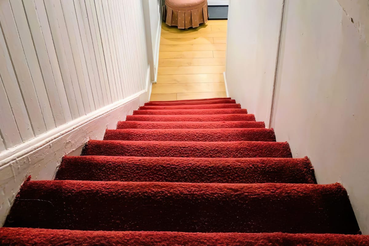 A staircase with red carpeted steps leading to a room with a wooden floor and a pink chair.
