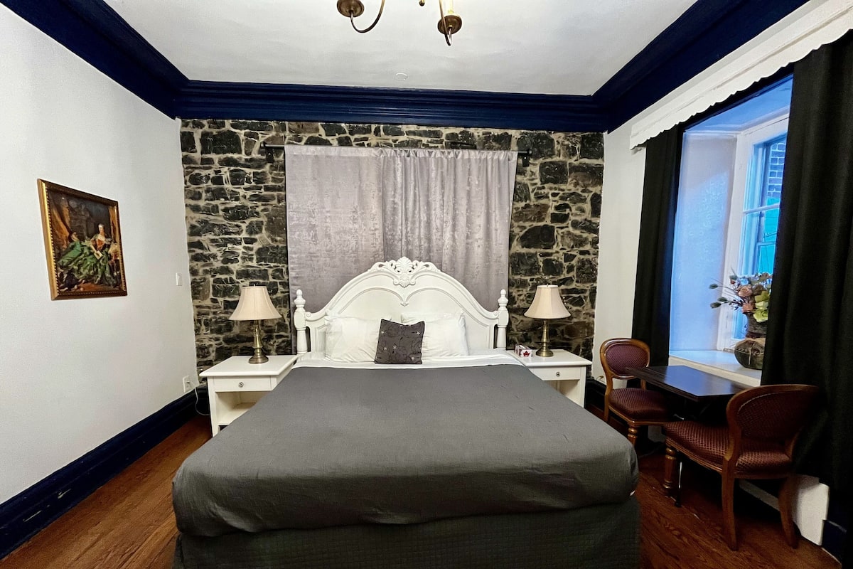 A bedroom with a stone wall, a large bed with a white headboard, two bedside tables with lamps, a chair, and a window with curtains.