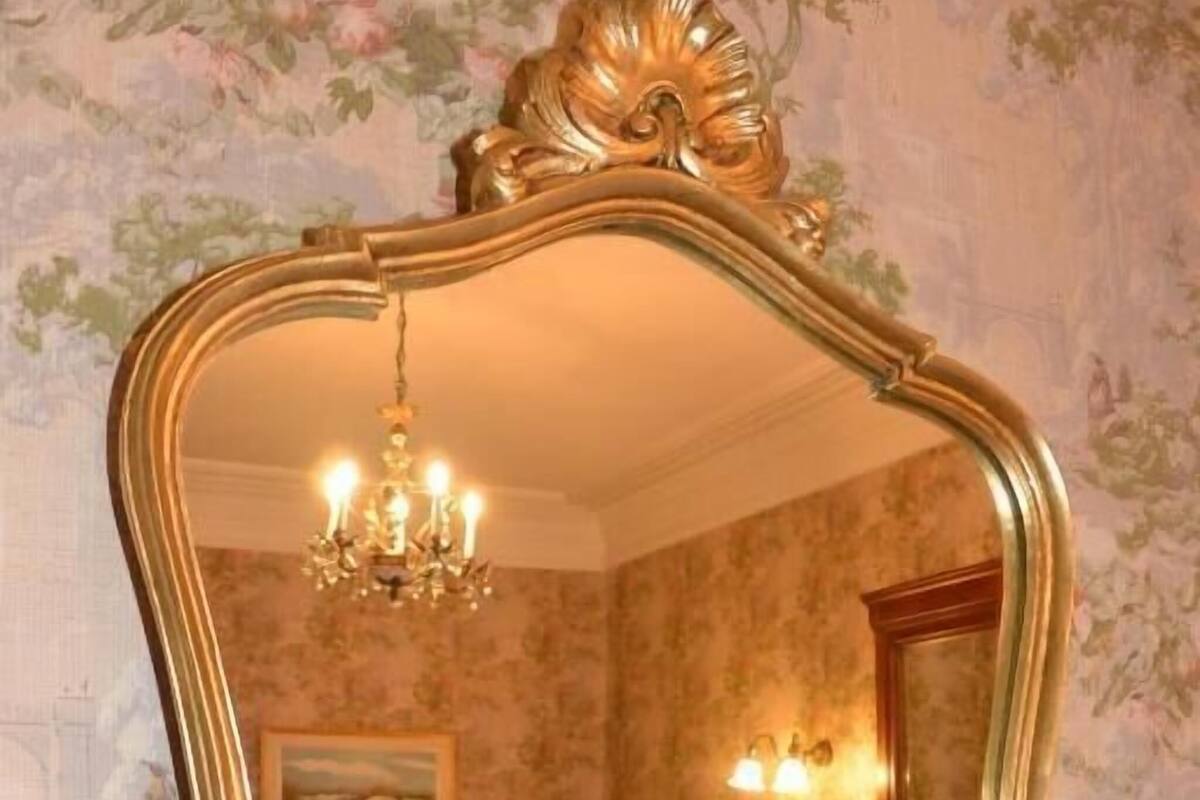 A bedroom with a large ornate mirror, a chandelier, a bed, and a fireplace.