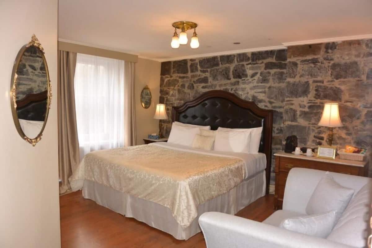A bedroom with a bed, a chair, a nightstand, a lamp, and a stone wall.