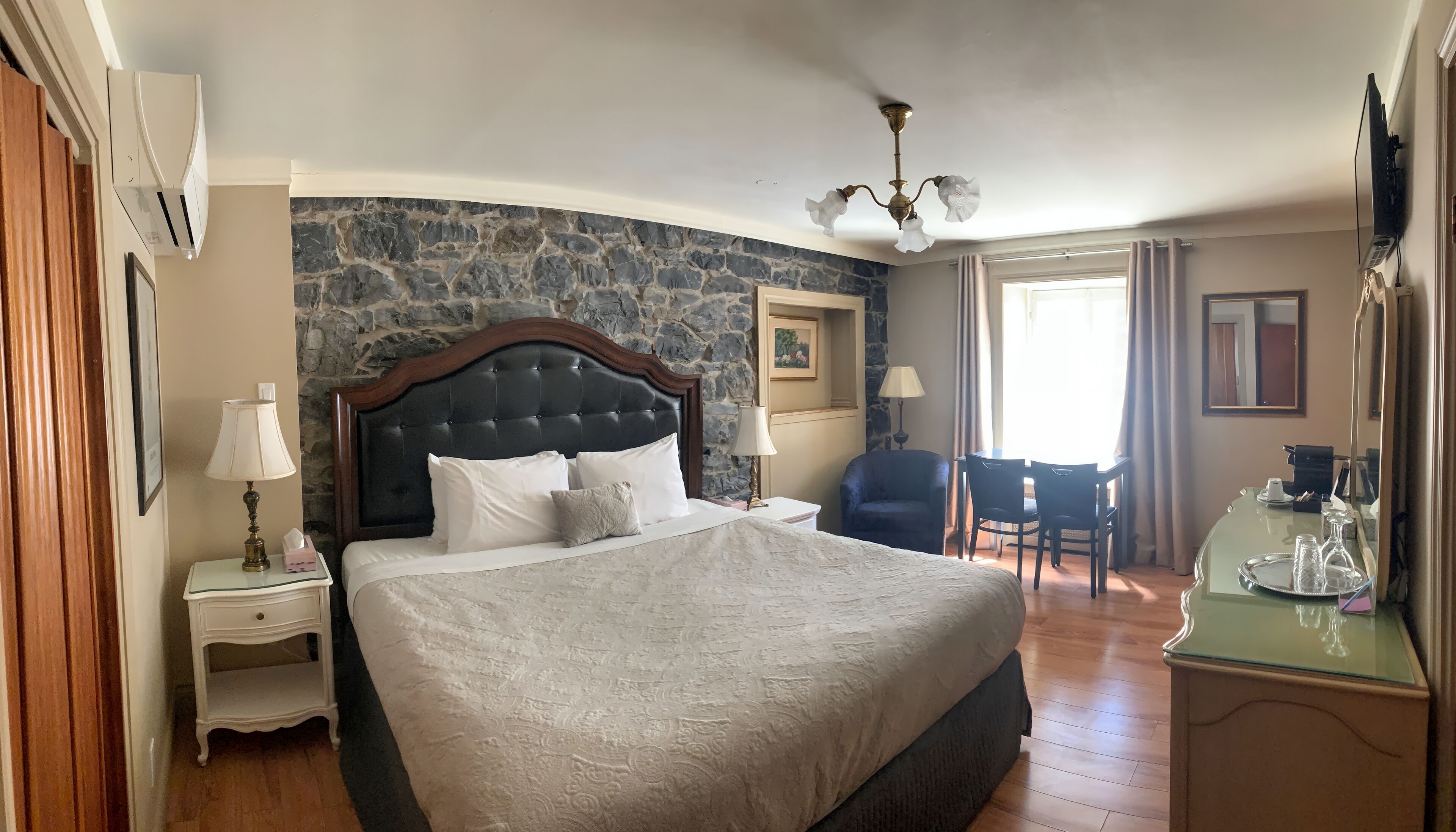 Habitación de hotel con una cama grande, un escritorio con silla, una mesita de noche con lámpara y una pared de piedra.