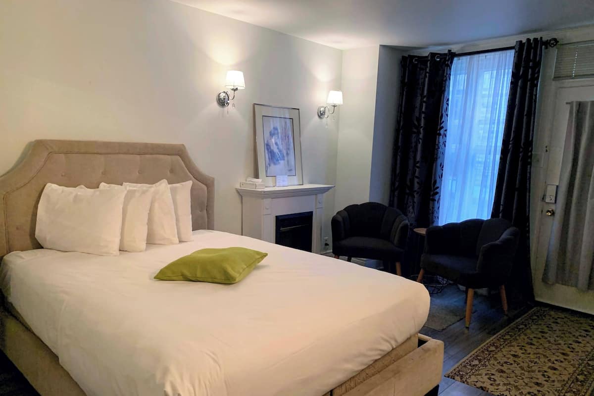 A hotel room with a bed, a fireplace, a desk, two chairs, and a view of the outside through a window with curtains.