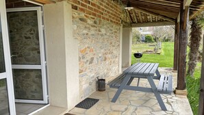 Un patio couvert avec un mur en pierre, une table en bois et un barbecue à l’arrière-plan.