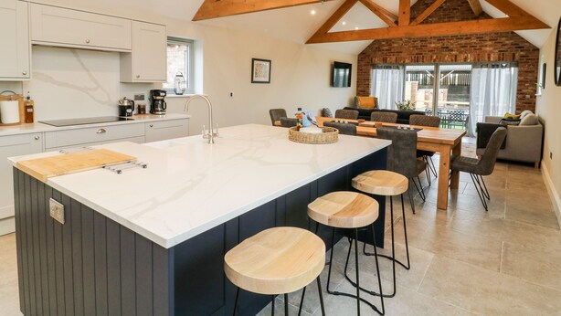 A modern kitchen with a large island, wooden beams, and a dining area with a brick wall. - The Cart House (York)