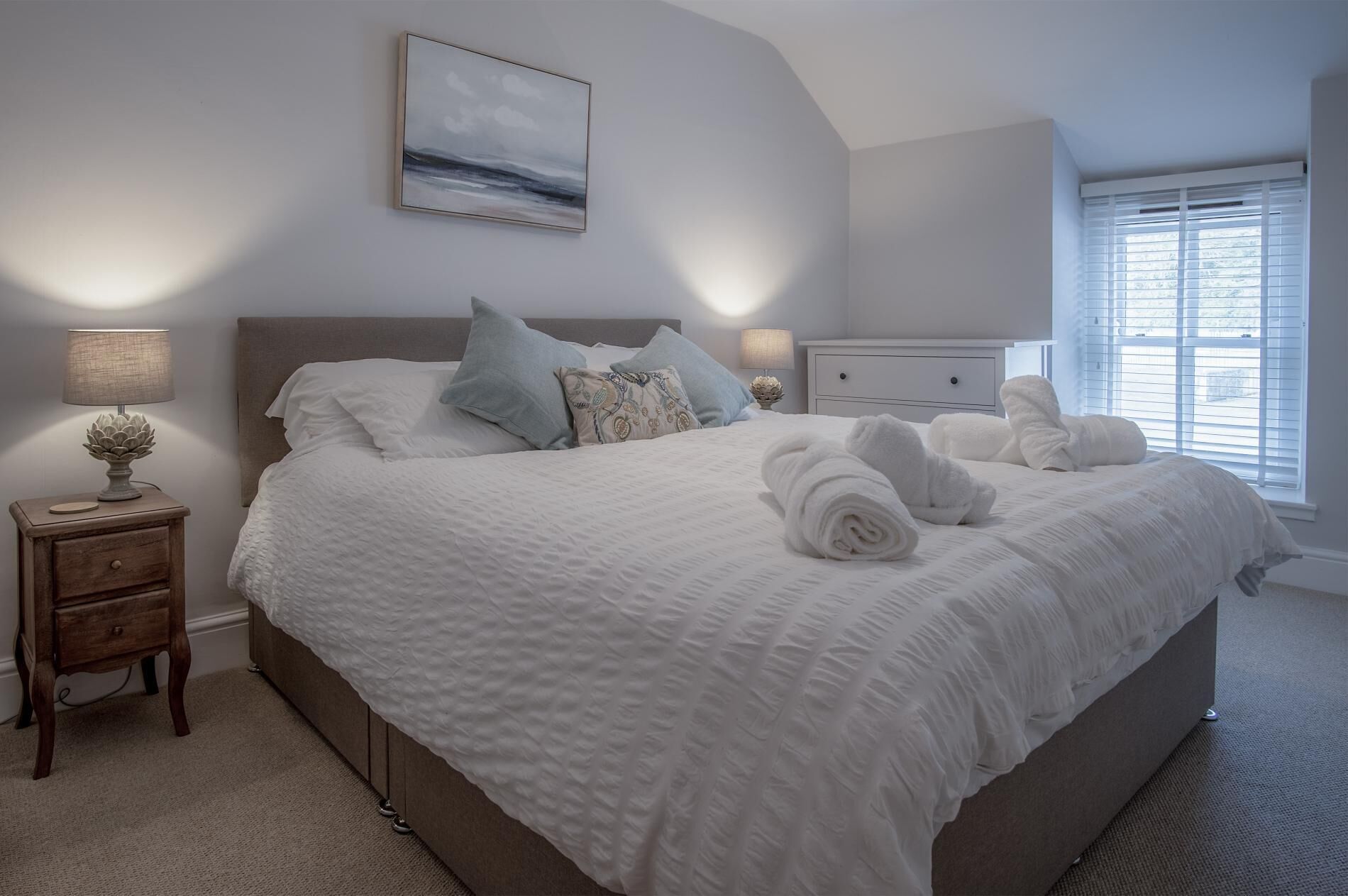 A bedroom with a large bed, a nightstand, a lamp, a bedside table, and a window with blinds.