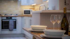 A modern kitchen with white cabinets, a microwave, and a wine bottle with glasses on the counter. - Noe Acre - 3 Bedroom Cottage - Tenby (Tenby)