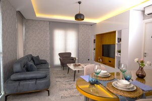 Una sala de estar moderna con un sofá gris, una mesita con un jarrón y una copa de vino, y un televisor en la pared.