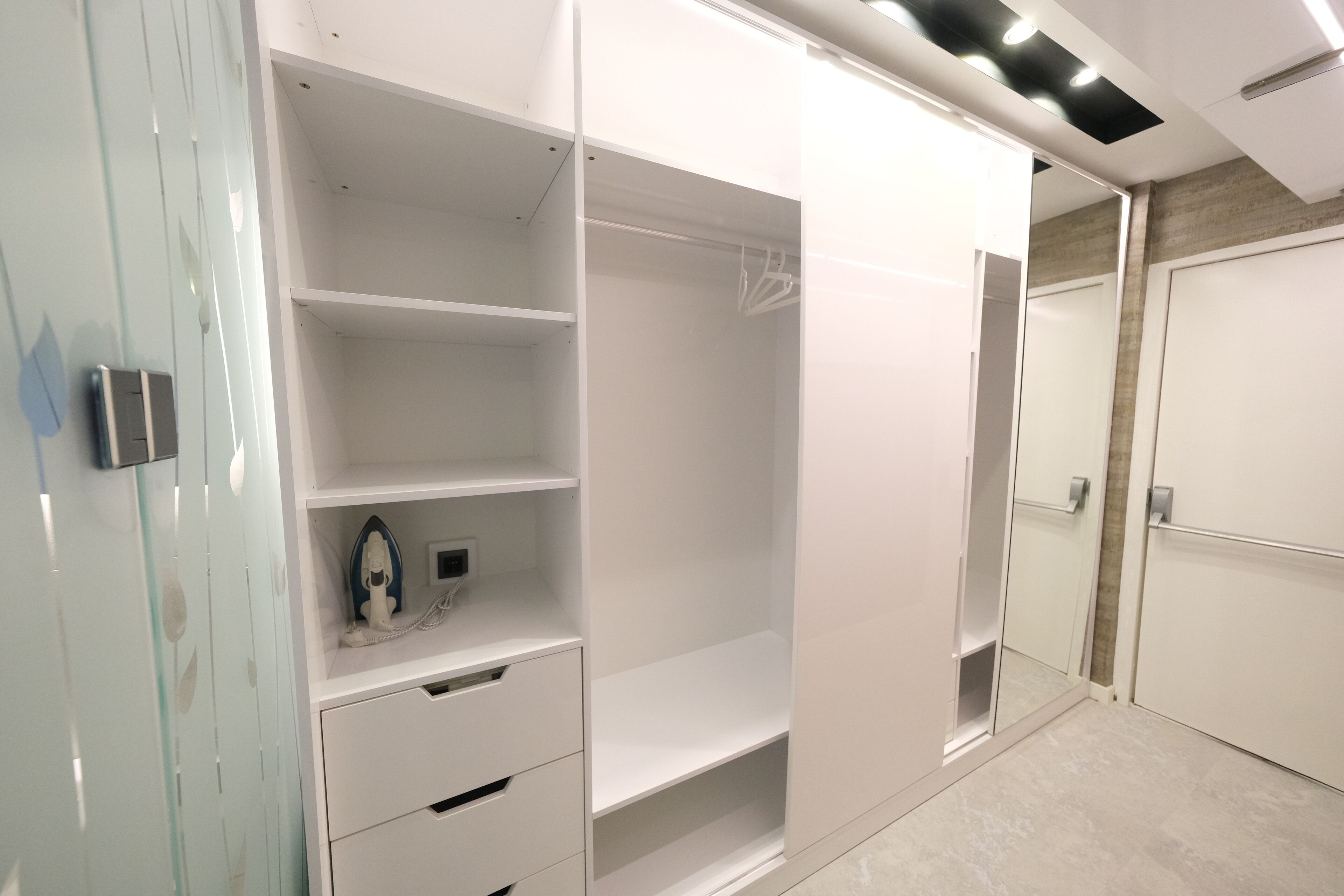 A modern, white walk-in closet with multiple shelves and drawers, a small ironing board, and a mirror.