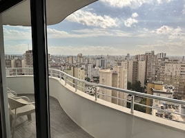 Un balcón con vista a la ciudad, barandilla de metal y una silla.
