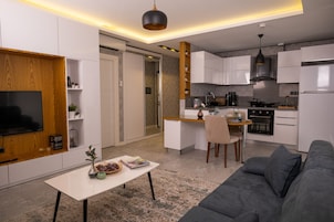 Una cocina moderna con armarios blancos, un comedor con mesa y sillas, y un sofá gris en la sala de estar.