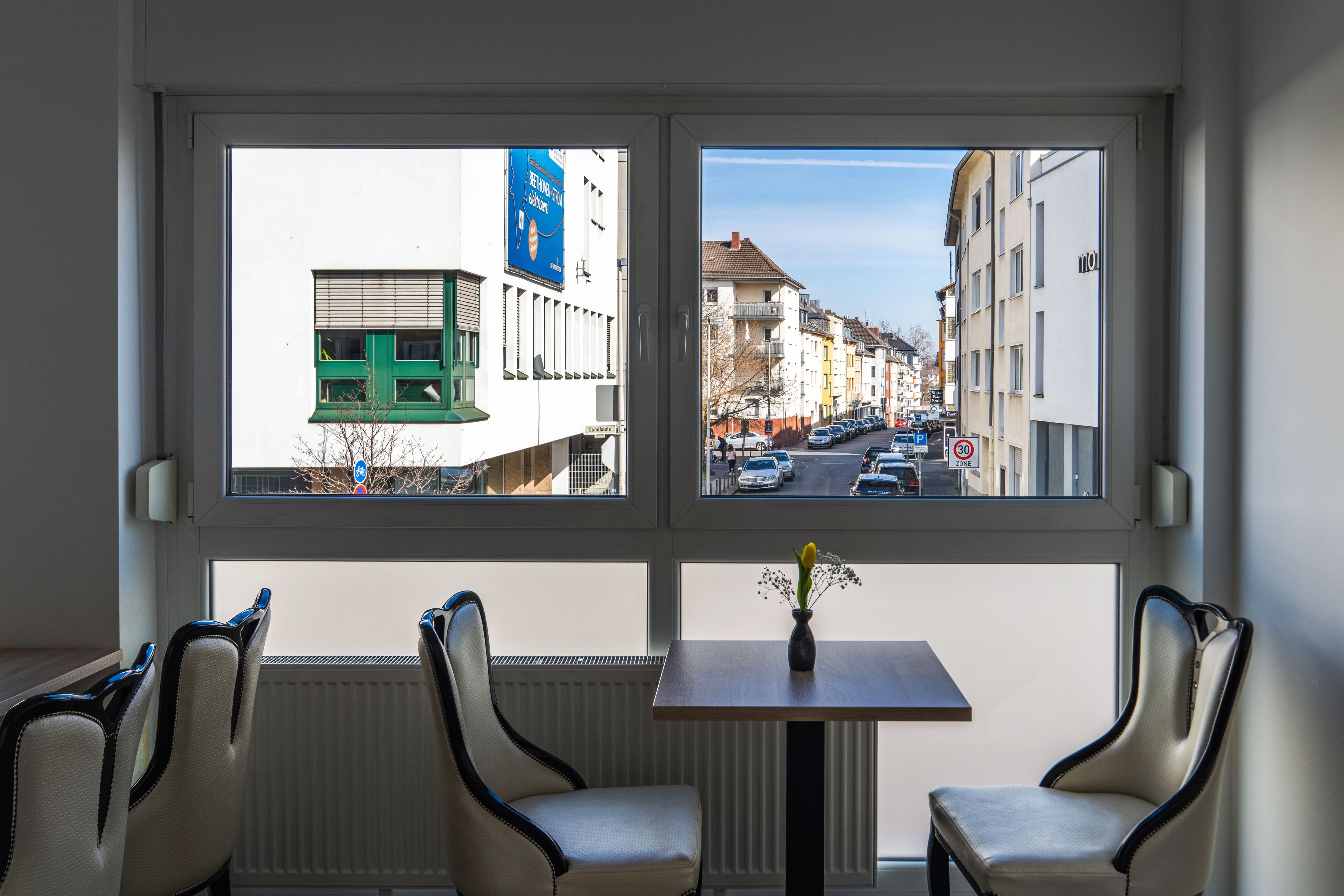 A hotel room with a large window offering a view of a city street. There are two chairs and a small table with a vase of flowers.