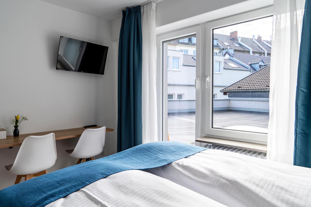 A hotel room with a bed, a desk with two chairs, a TV mounted on the wall, and a window with curtains.