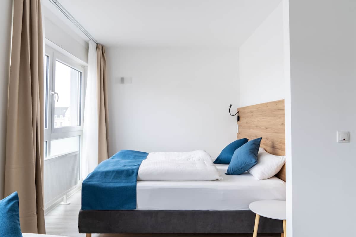 A modern hotel room with a bed, a small round table, and a view of the outside through a large window.