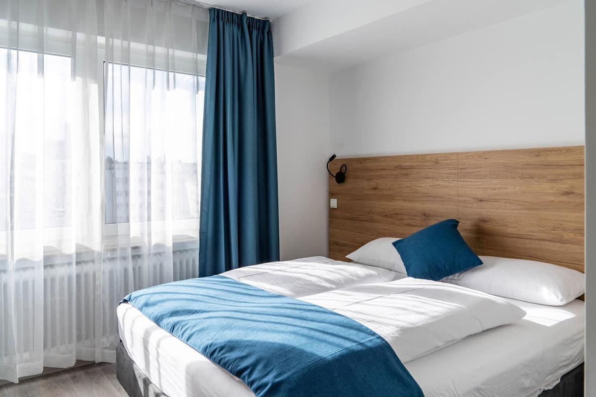 A hotel room with a bed, a wooden headboard, a window with sheer curtains, and a blue accent pillow.