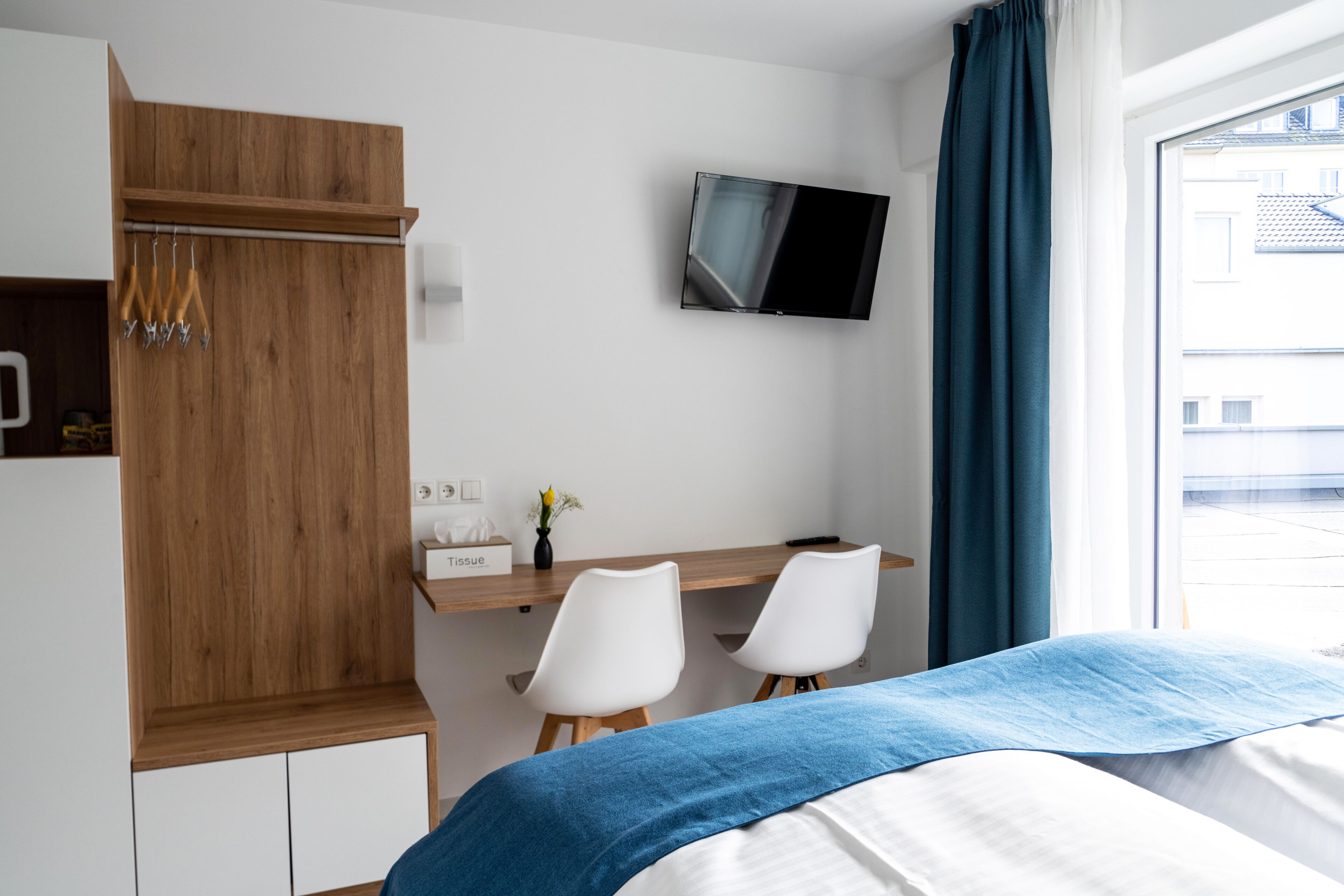 A hotel room with a bed, a desk, two chairs, a TV, and a window with curtains.
